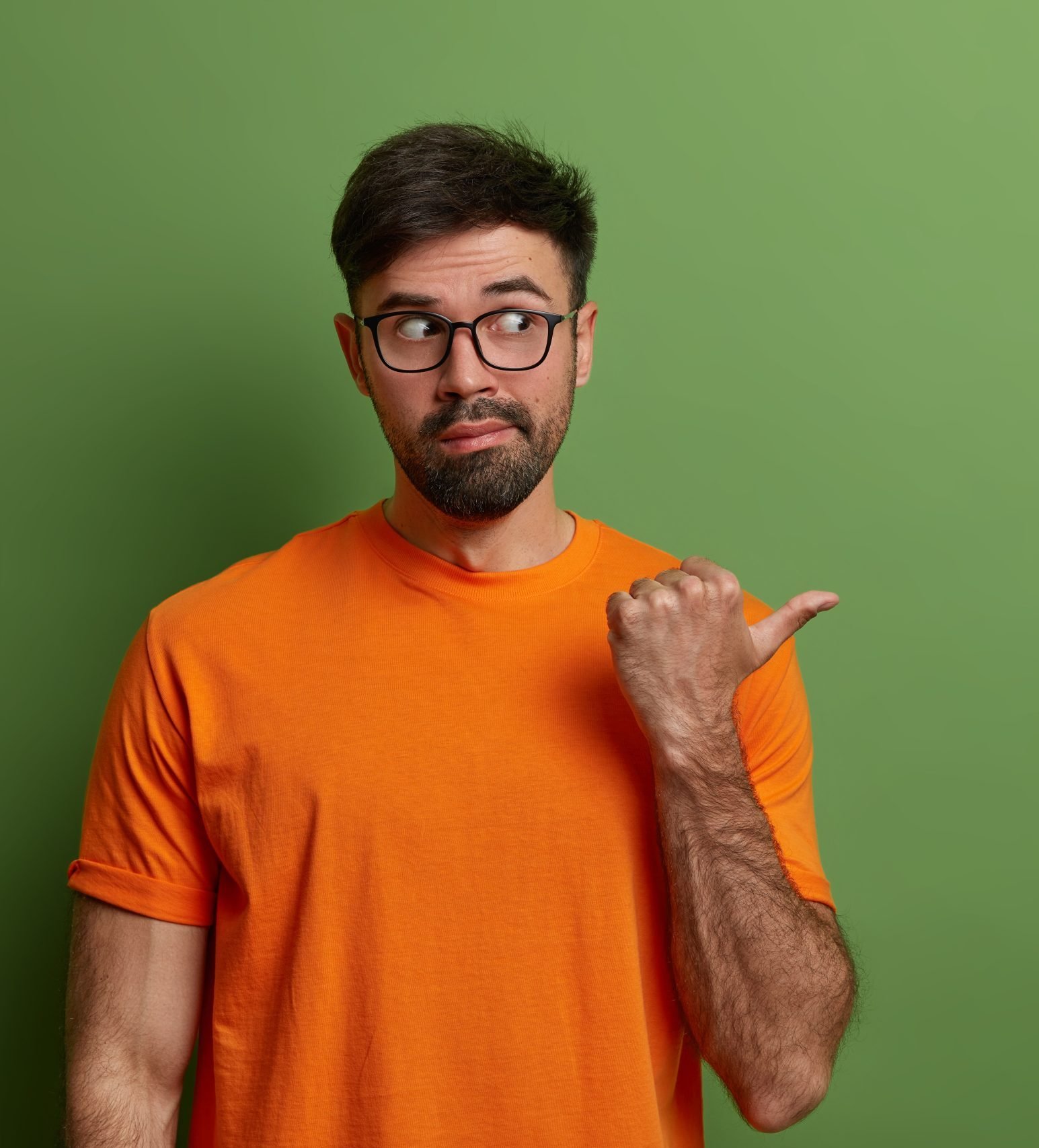 Curious man looks with suspicion and interest aside, points aside on blank space, shows advertisement of product or company banner, gestures against green background, wears glasses, t shirt.